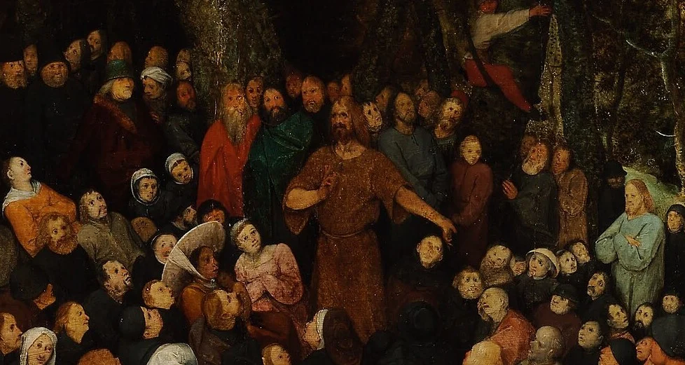 The preacher is small and far away. His arm points sideways.