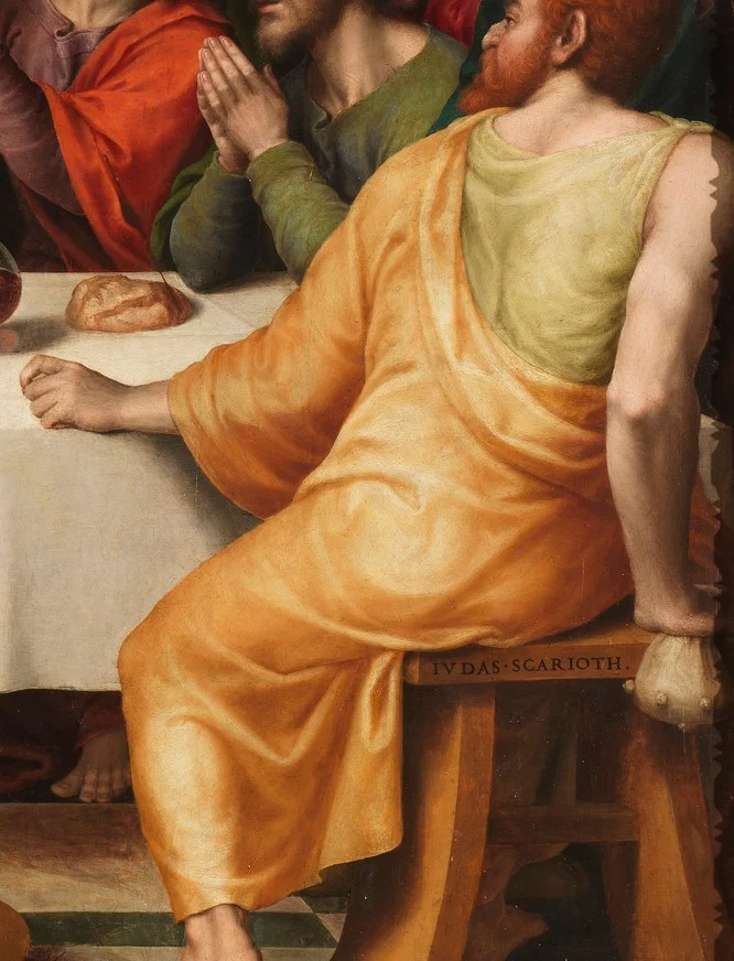 Judas wendet sich vom Tisch ab. Der Beutel ist hinter seinem Rücken, und sein Name, Judas Iskariot, steht auf dem Schemel.